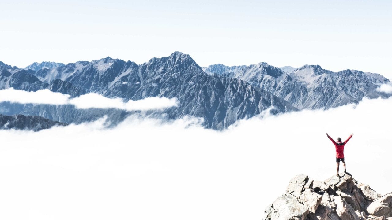 A man in red standing on a mountain above the clouds with his arms in the air.