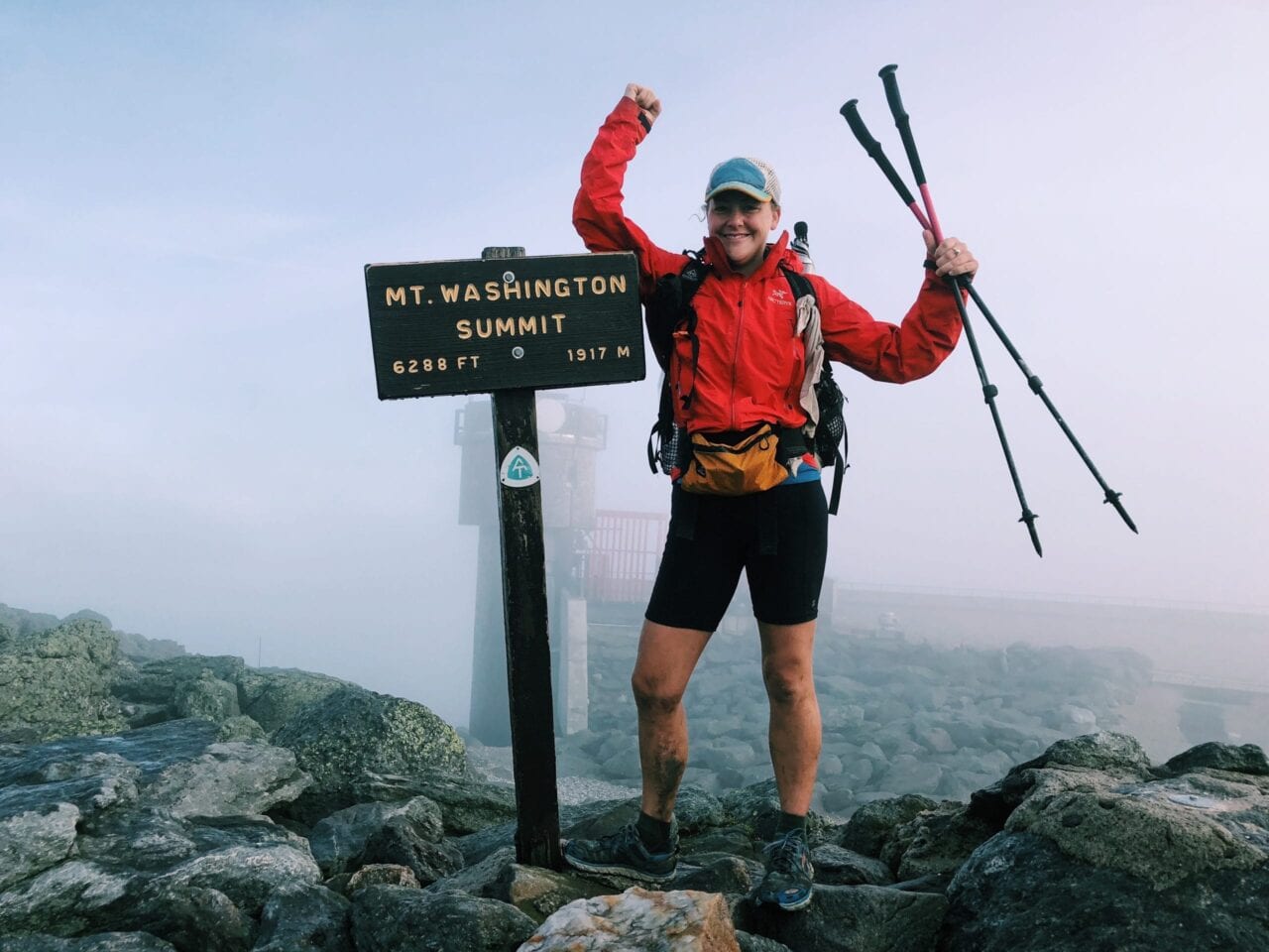 A woman hiker standing next to the Mt Washington Summit sign, holding her trekking poles up.