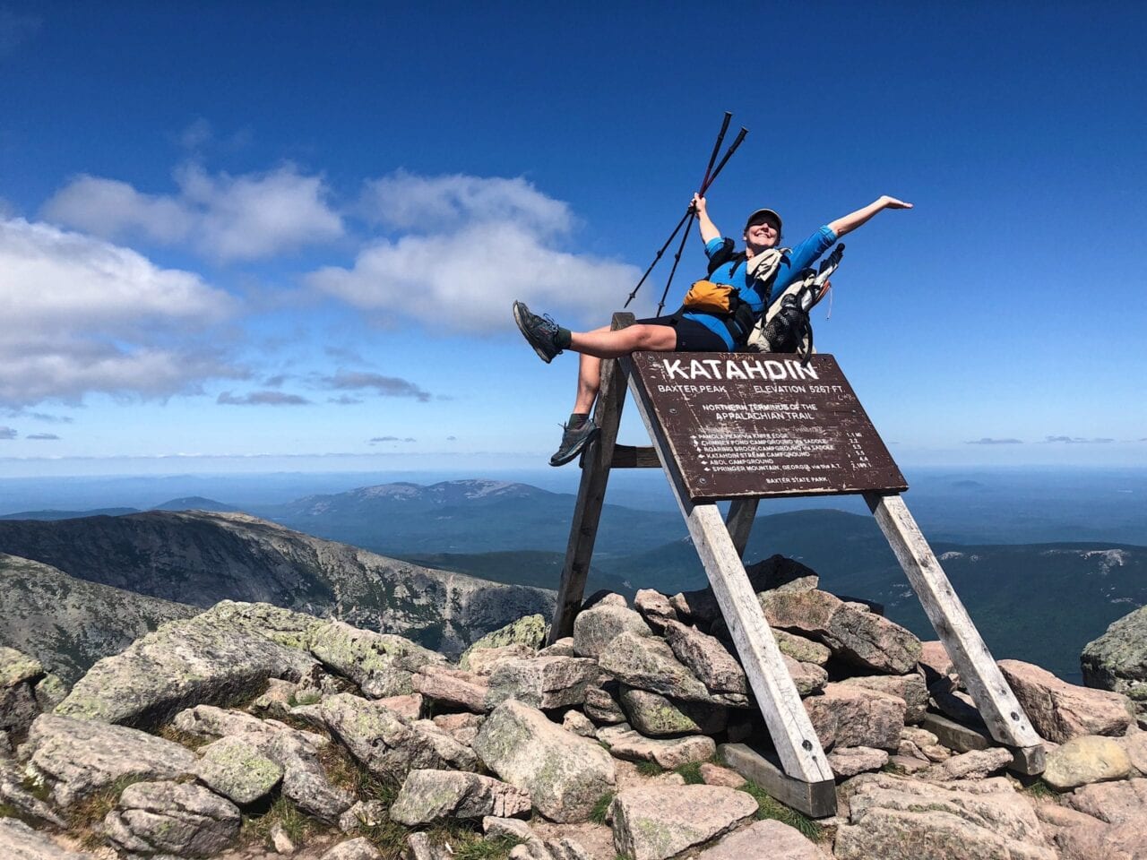 A woman sitting on the Mt Katahdin sign with her arms and leg in the air.