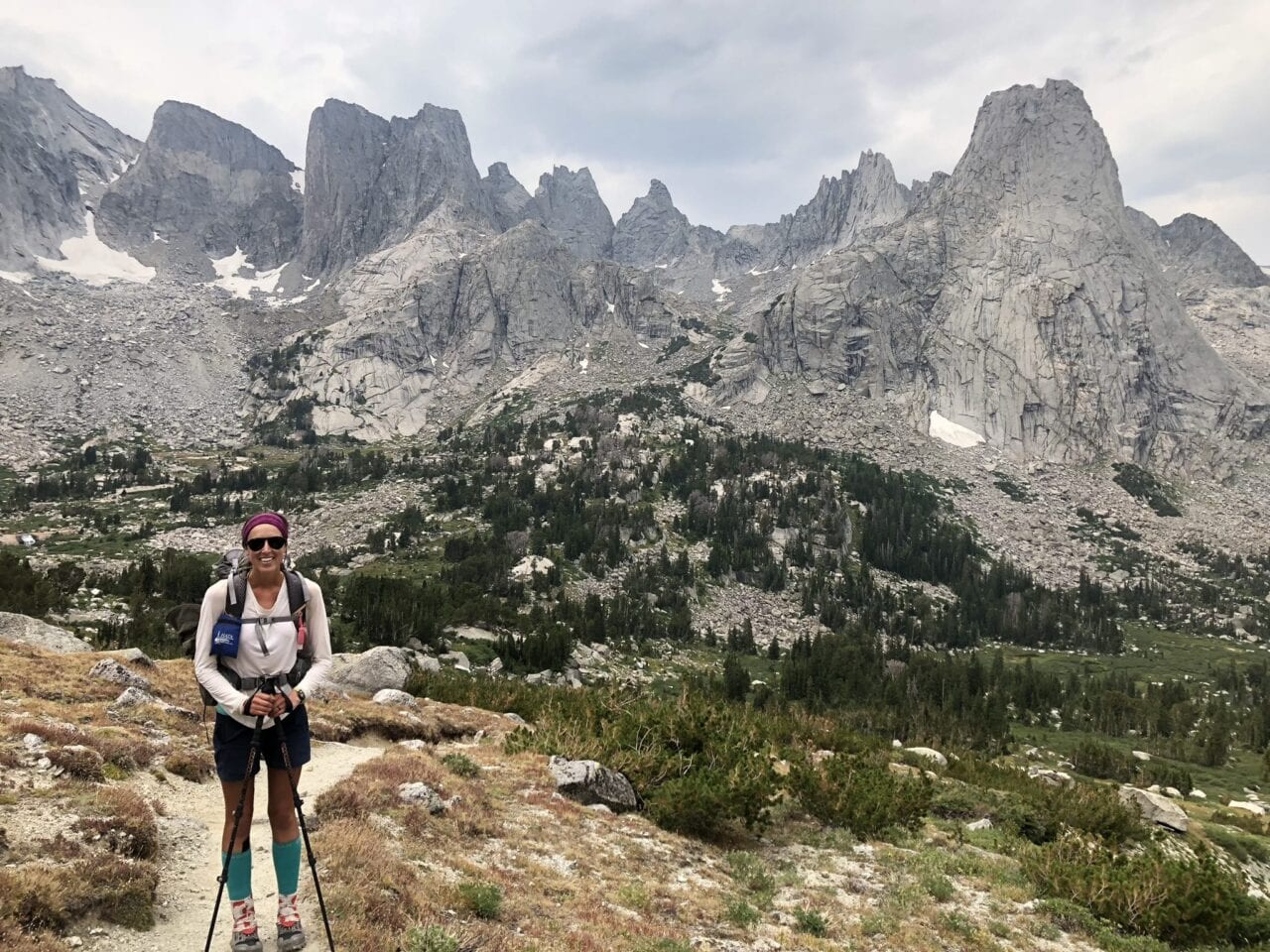 A hiker stands on a trail with beautiful mountains in the background.