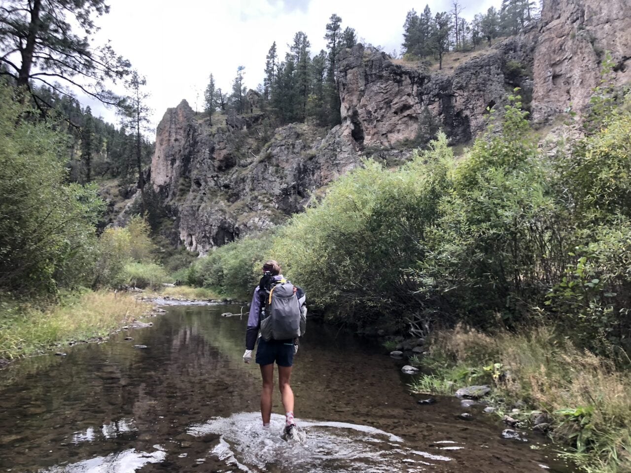 A hiker walking in a river with trees, bushes, and mountains on both sides.