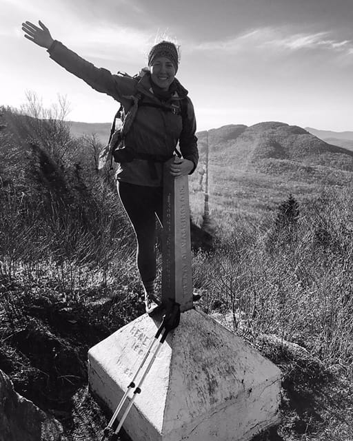A hiker stands on a trail terminus with her arm in the air.