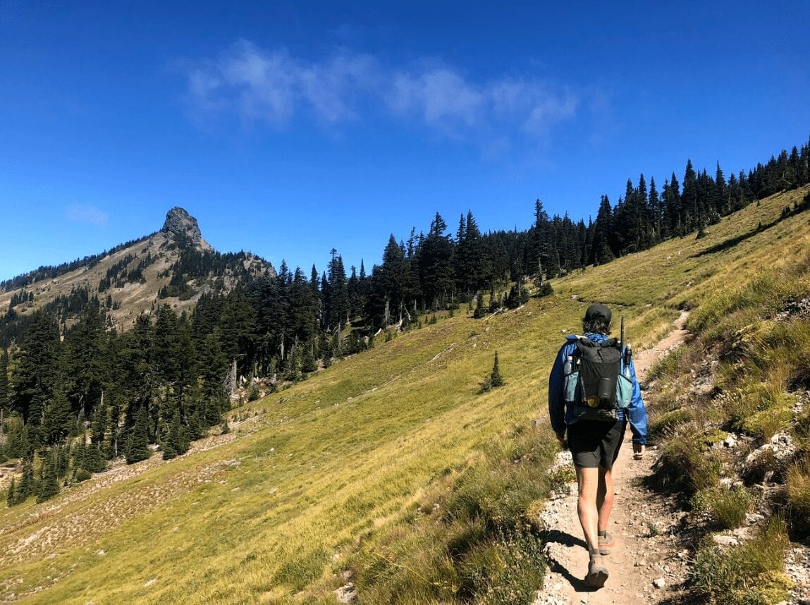 A hiker walking on a trail on the side of a green mountain hillside.