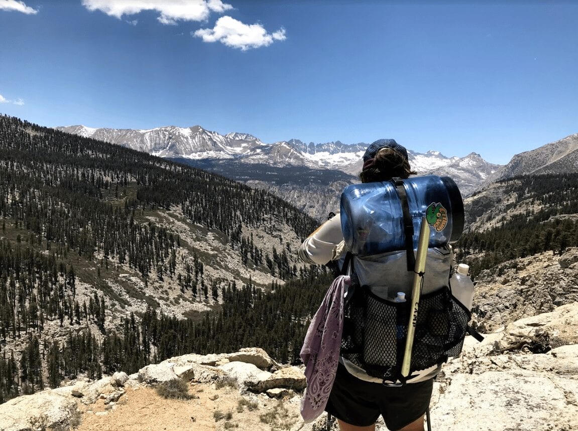 A hiker stands with her backpack and bear canister strapped to her back as she looks over the mountains.
