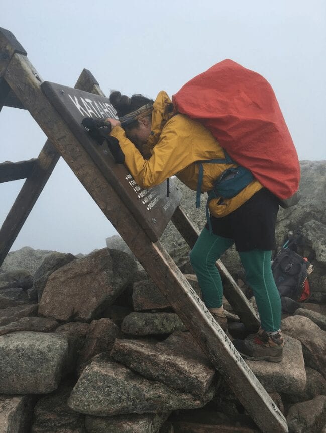 A hiker resting her head against the Mount Katahdin sign with her backpack on.