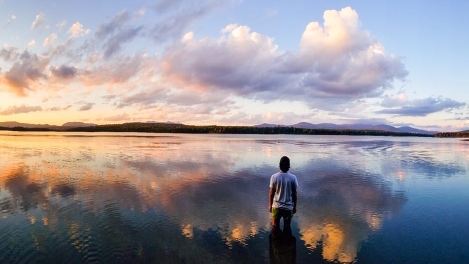 A man stands in front of a lake with a sunset reflecting in the water.