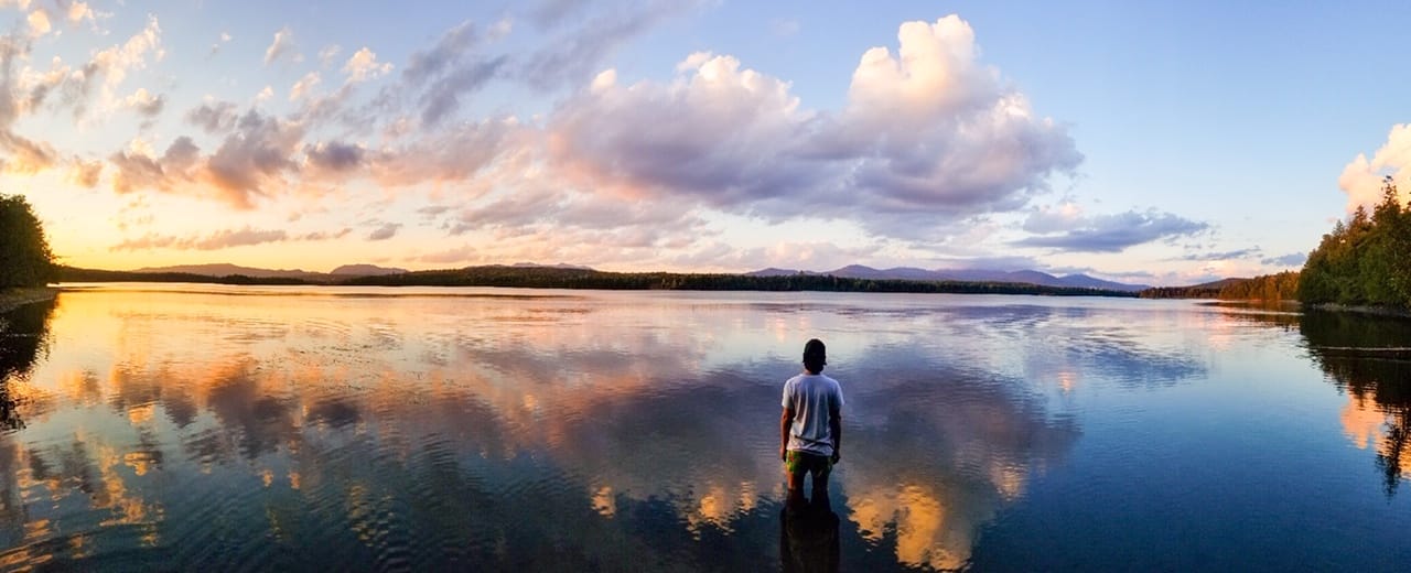 A man stands in front of a lake with a sunset reflecting in the water.