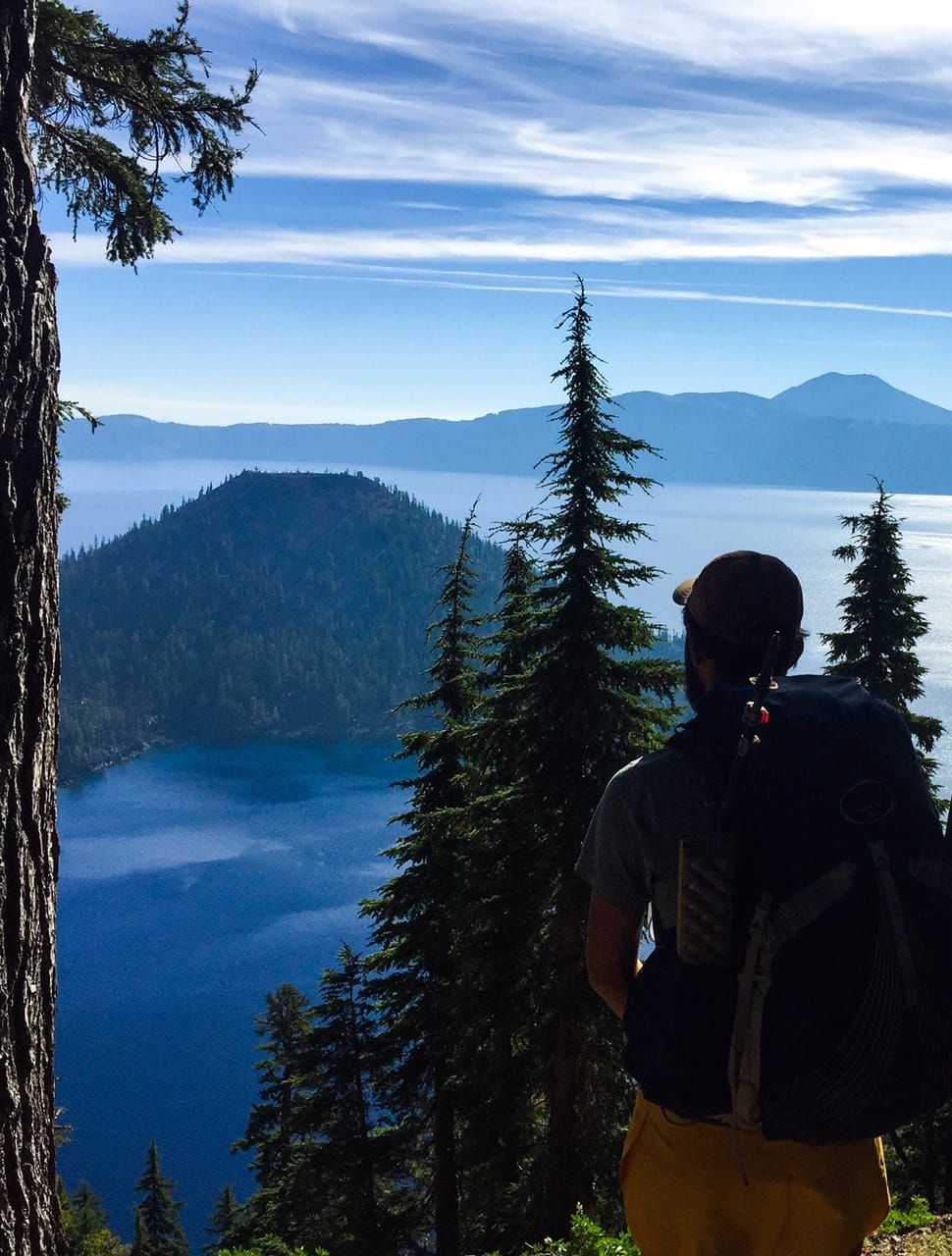 A hiker standing with his backpack on looking out at a mountain and lake below.