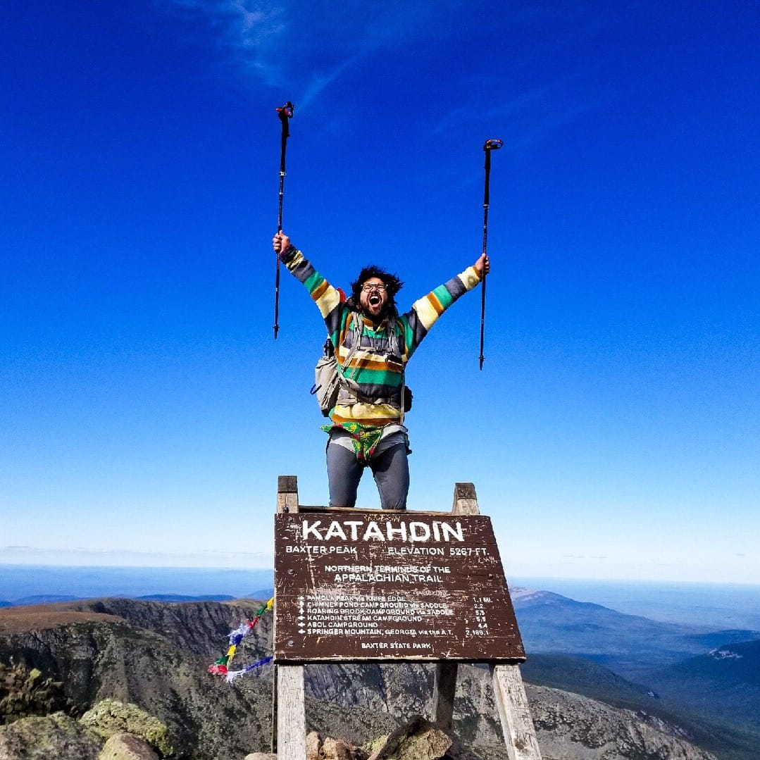 A hiker standing on the sign on top of Mount Katahdin holding his trekking poles in the air.