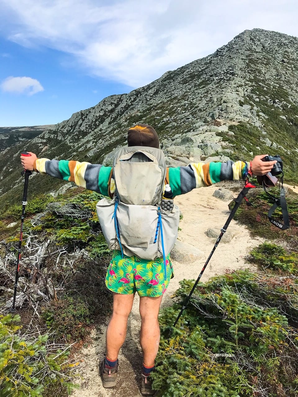 A hiker standing on a trail with his arms out holding trekking poles.