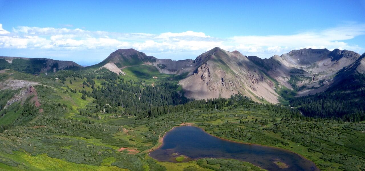 A blue lake sits in a green meadow in front of impressive rocky mountains.