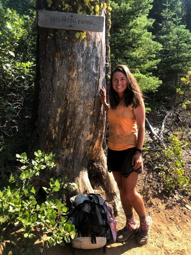 A woman stands next to a tree with a trail sign on it.