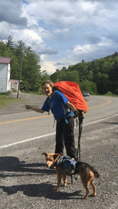 A woman hitch-hiking with her dog on the side of the road.