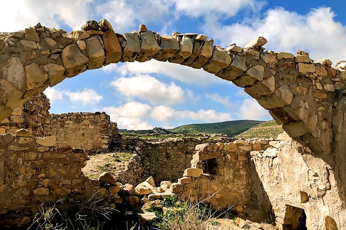 An arch and some ruins sit in a green field with distant rolling hills and a blue sky in the background.
