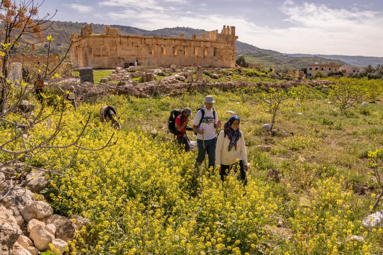 A group of hikers walk through a desert field in front of a castle.