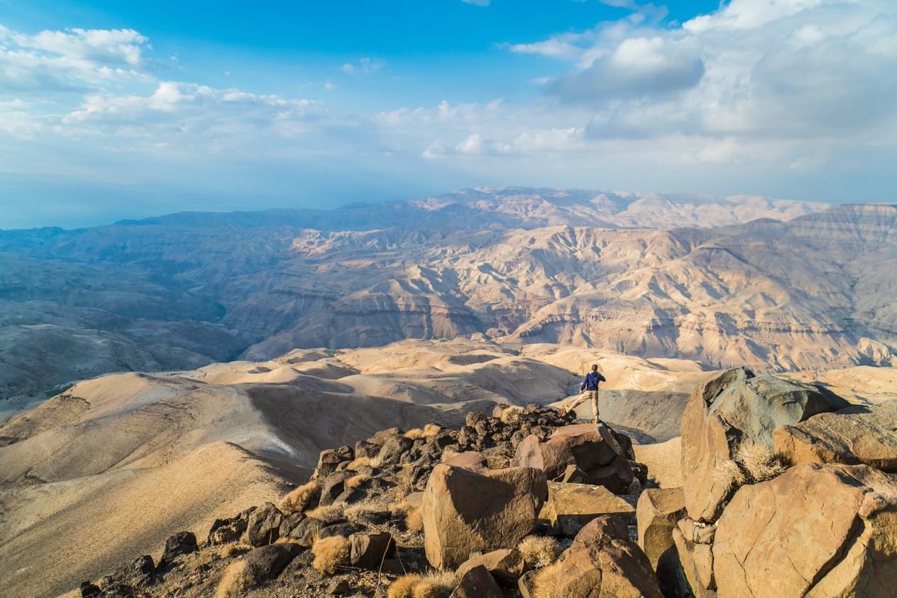 A hiker stands on top of tan rocks against a backdrop of mountains.