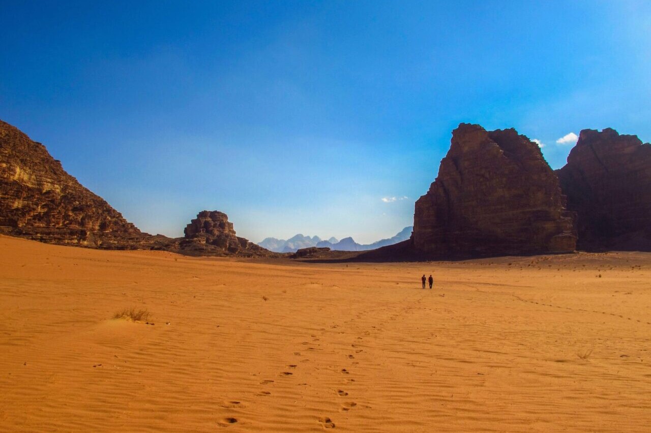 Two hikers walk through a red sandy desert towards distant rock formations.