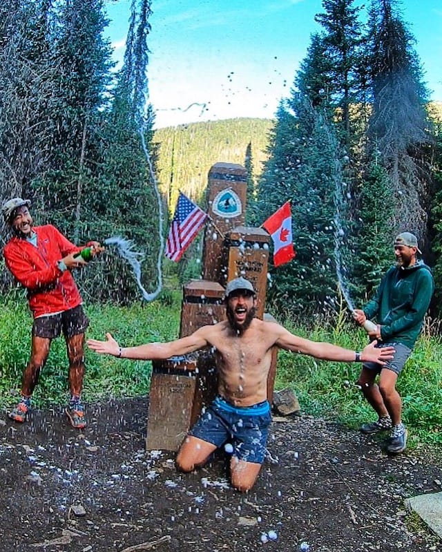 Three Pacific Crest Trail thru-hikers celebrating and popping champagne at the northern terminus.