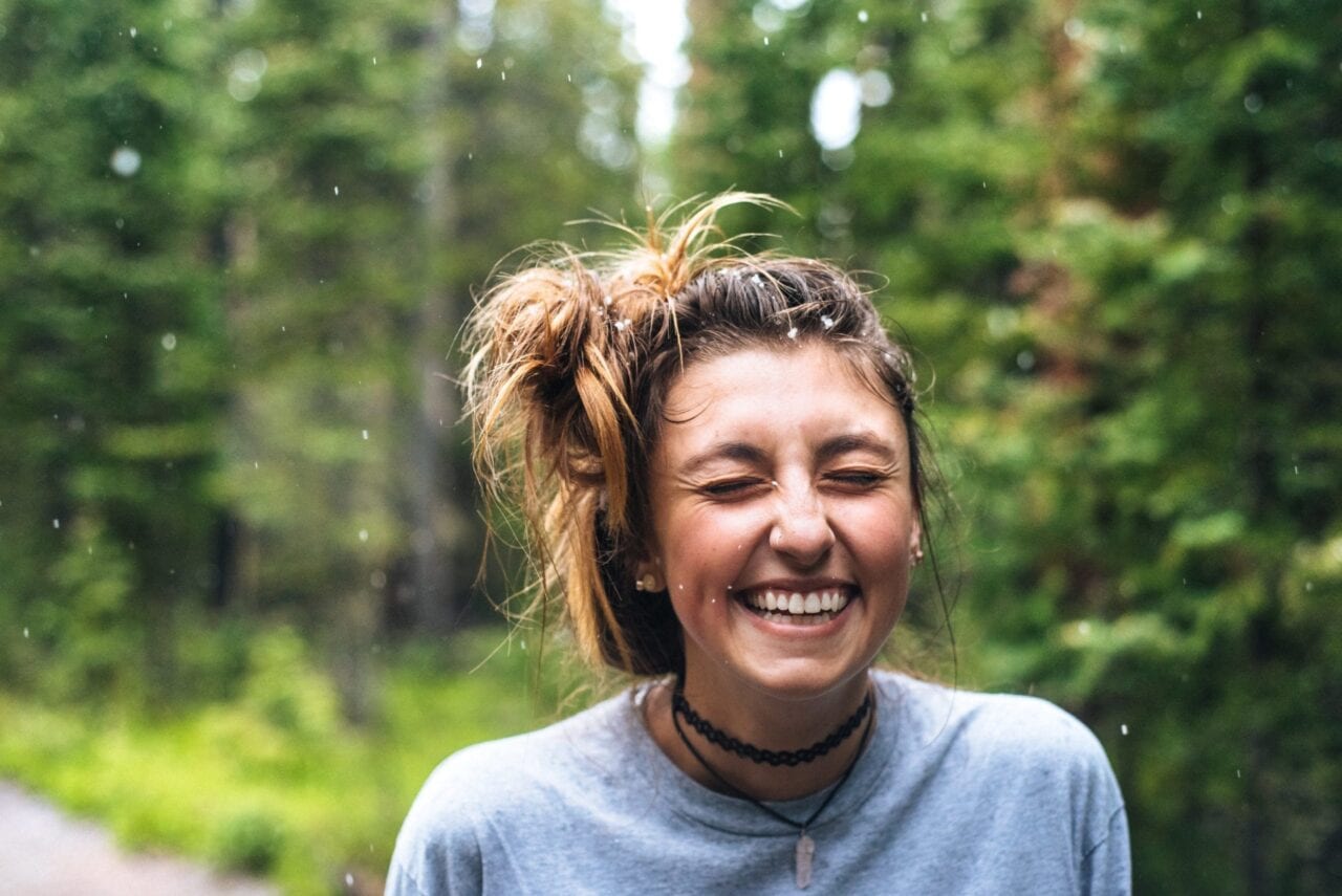 A girl with borwn hair smiles in the rainy green forest.