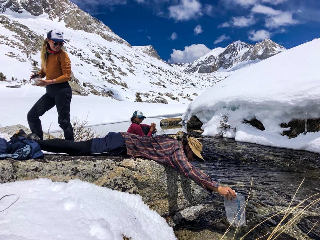 A hiker collecting water from a snowy creek.