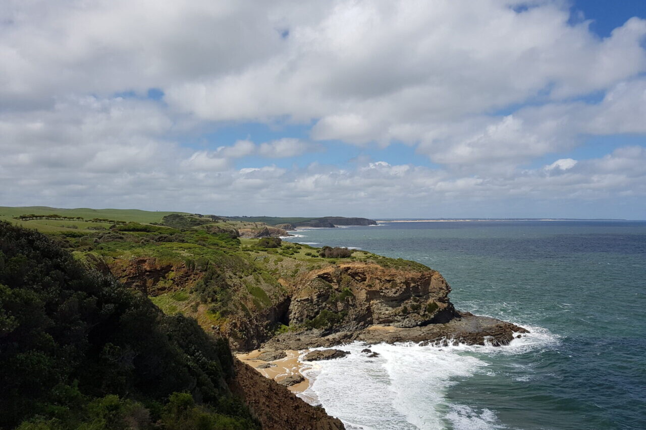 Ocean waves lap a sandy beach below rocky cliffs and green headlands.