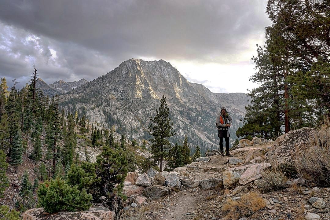 A hiker standing on a trail with a mountain in the background.