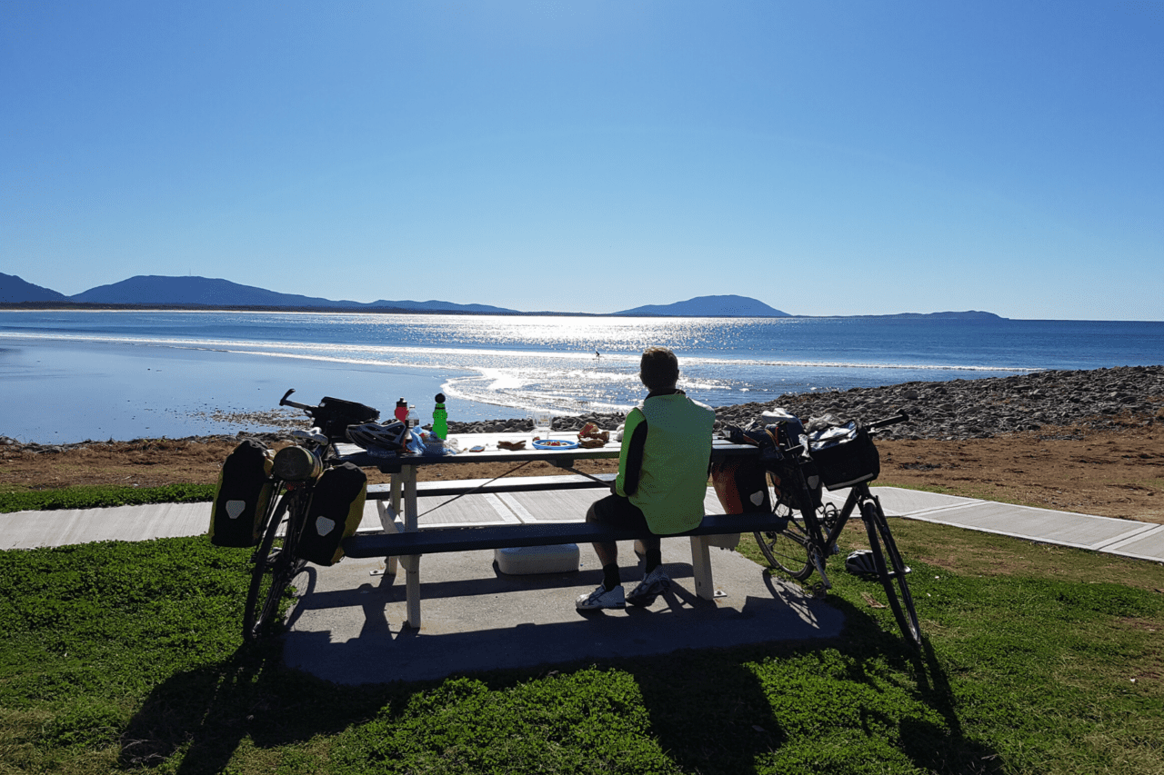 A person sitting at a picnic table next to a bike looking out at the ocean.