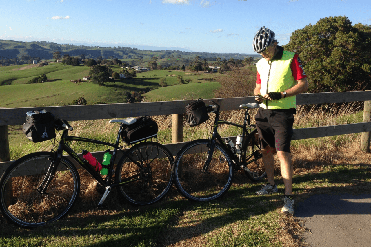 A man standing next to his bike along a fence with green hills and trees in the background.