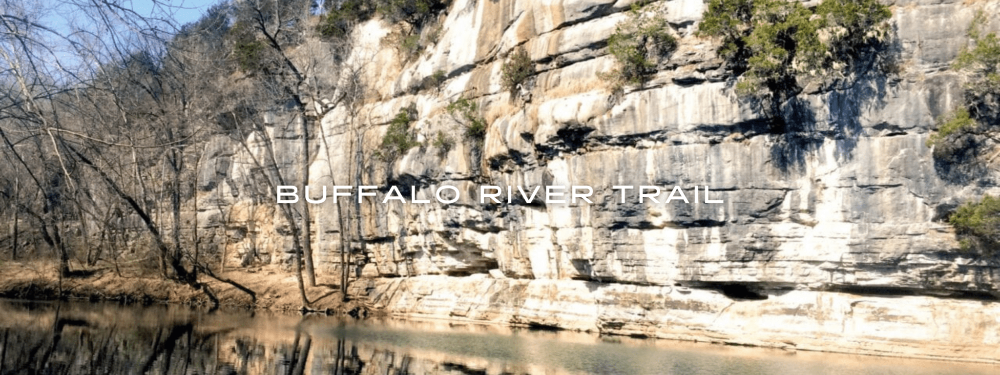 A glassy calm river flows by a tall rock wall under blue skies.