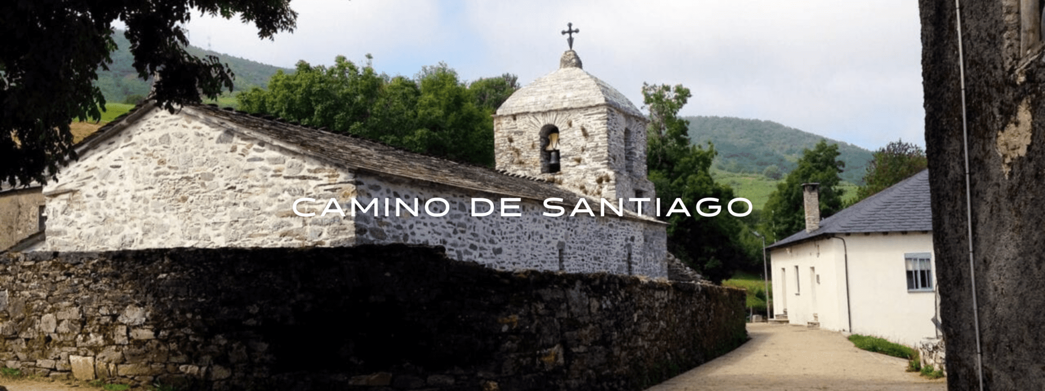 The Camino de Santiago winds through a small town on cobblestone streets with an old, white stone church on the left and green rolling hills in the background.