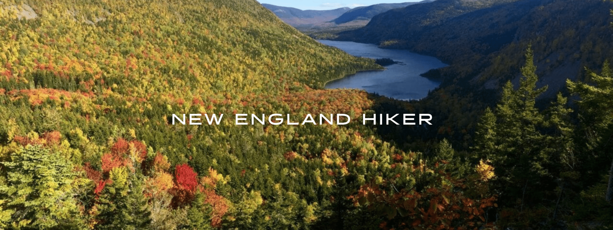 A view from a mountain ridge overlooks orange, red, yellow, and green trees and a blue lake in Baxter State Park in Maine.
