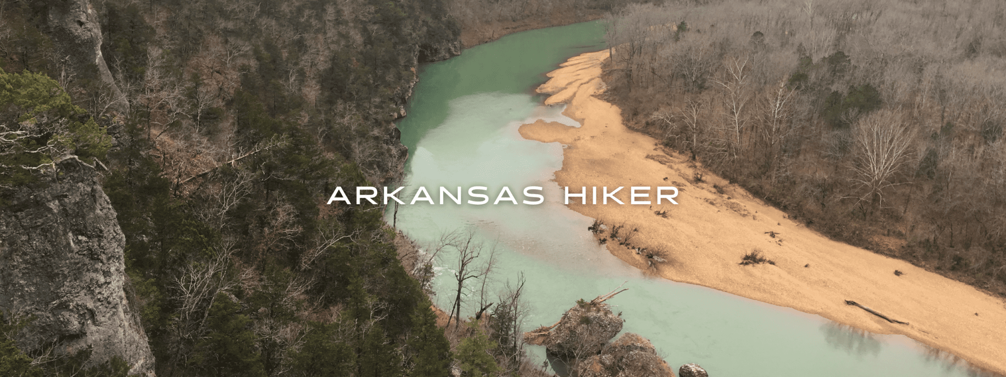 On the Ozark Highlands Trail in Arkansas a view looks over a teal river with a sandy beach and dense forests.